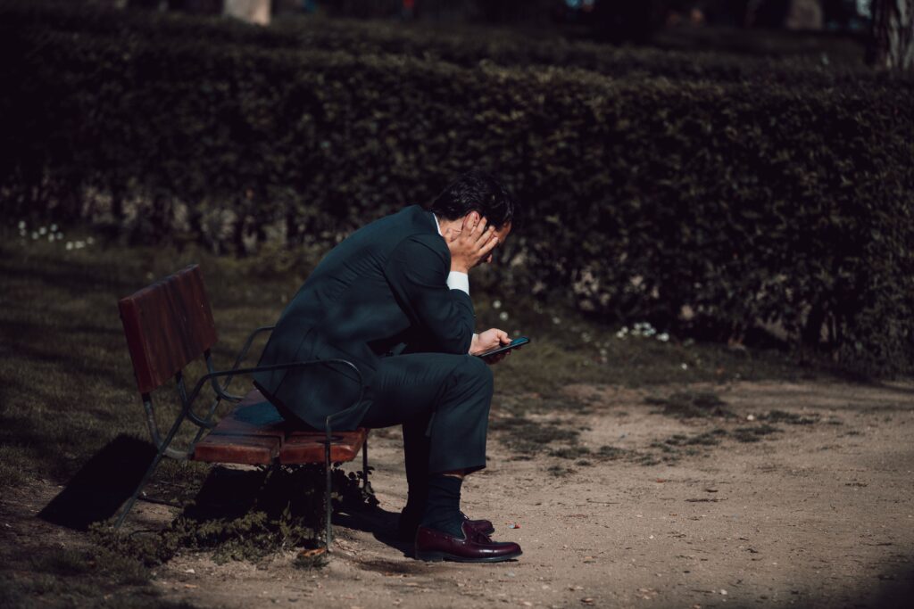 A worried businessman sits on a park bench in Madrid, holding a smartphone. Captured outdoors in natural light.